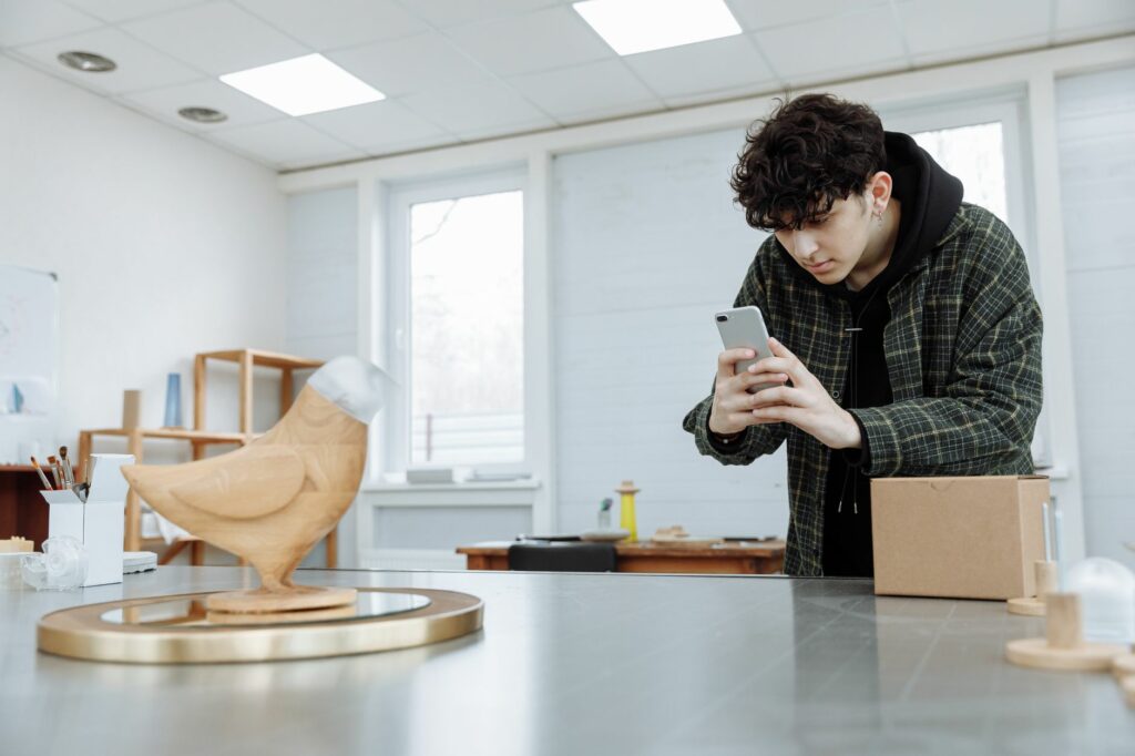 Homme en chemise à carreaux photographiant une sculpture en bois avec son smartphone dans une pièce très lumineuse