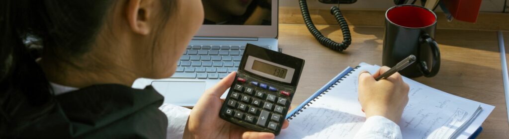 Une femme calcule un budget à son bureau, calculatrice en main, avec un ordinateur portable et un bloc-notes