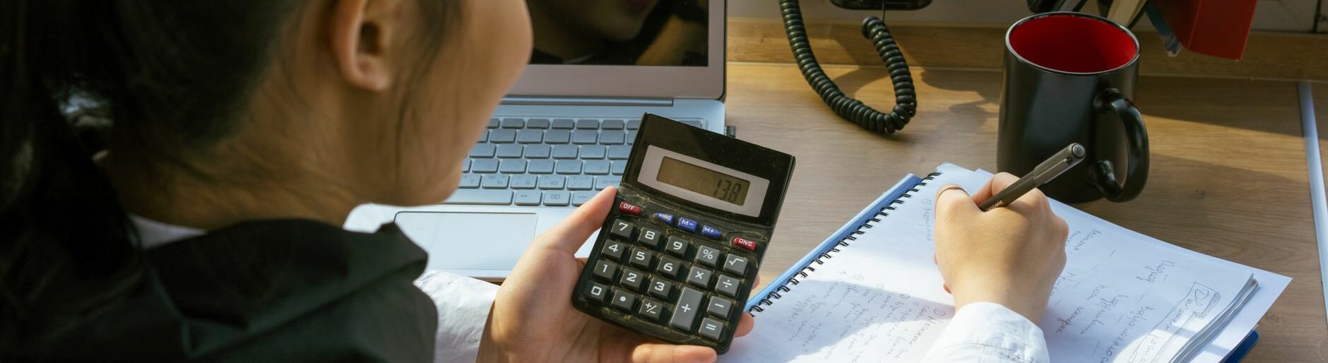 Une femme calcule un budget à son bureau, calculatrice en main, avec un ordinateur portable et un bloc-notes