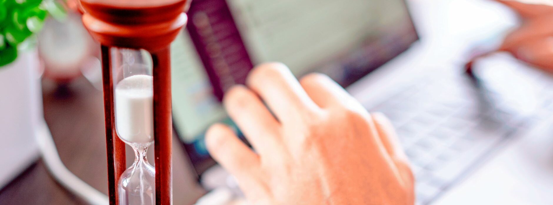 Un sablier posé à côté d’une main sur le clavier d’un ordinateur portable, sur un bureau en bois