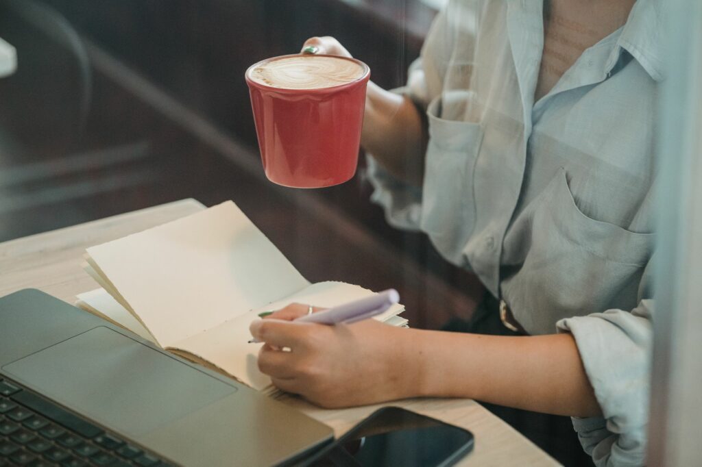Une femme écrit dans un carnet devant son ordinateur portable, un café posé à côté, dans un café