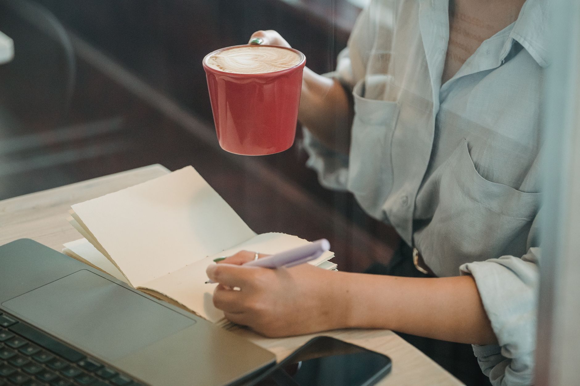 Une femme écrit dans un carnet devant son ordinateur portable, un café posé à côté, dans un café