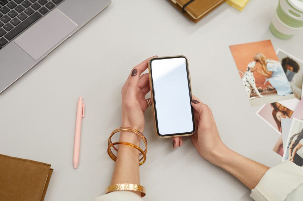 Gros plan sur une femme qui tient un smartphone, assise à un bureau avec un ordinateur portable et des photos imprimées