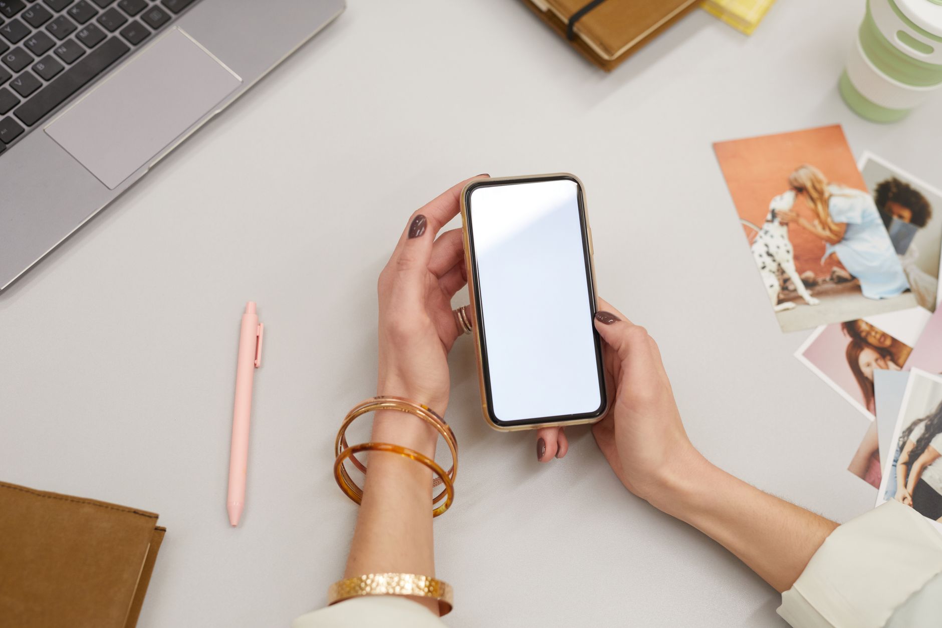 Gros plan sur une femme qui tient un smartphone, assise à un bureau avec un ordinateur portable et des photos imprimées